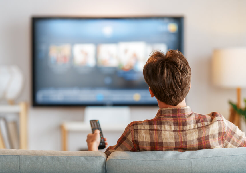 Mężczyzna siedzący na kanapie tyłem do obiektywu, trzymający w ręku pilota i przeglądający spersonalizowane rekomendacje filmów na dużym ekranie telewizora z menu platformy VOD.