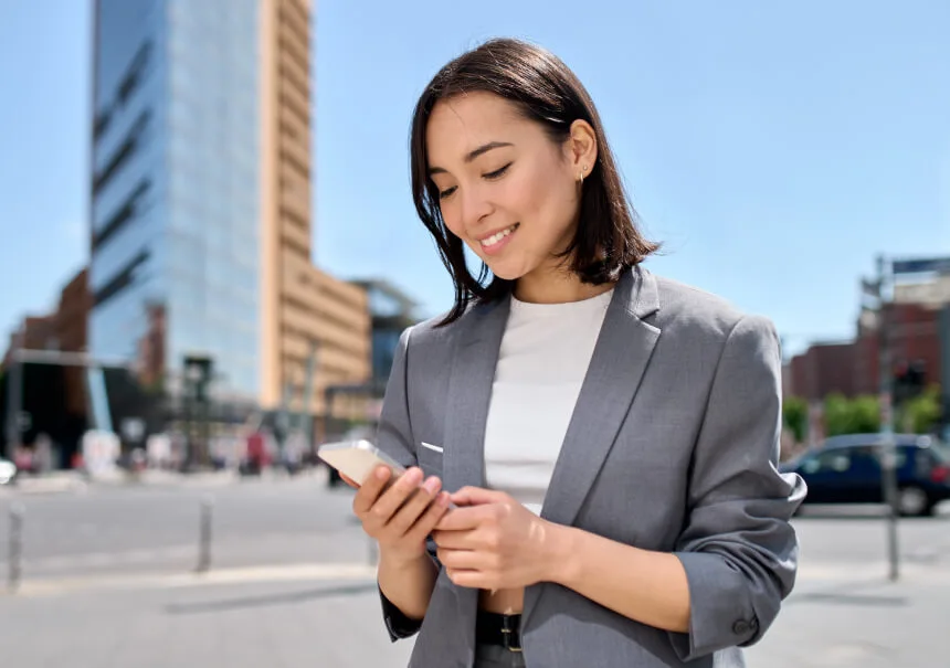 Klientka e-commerce w Polsce odbierająca wiadomości od sklepu internetowego na aplikacji WhatsApp. Uśmiechnięta kobieta w szarym żakiecie korzysta ze smartfona na tle nowoczesnej infrastruktury miejskiej.