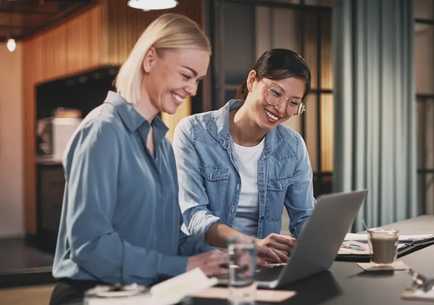 Dwie uśmiechnięte specjalistki ds. marketingu projektują stronę docelową w kreatorze landing page bez kodowania. Kobiety pracują przy laptopie w nowoczesnym biurze agencji interaktywnej.