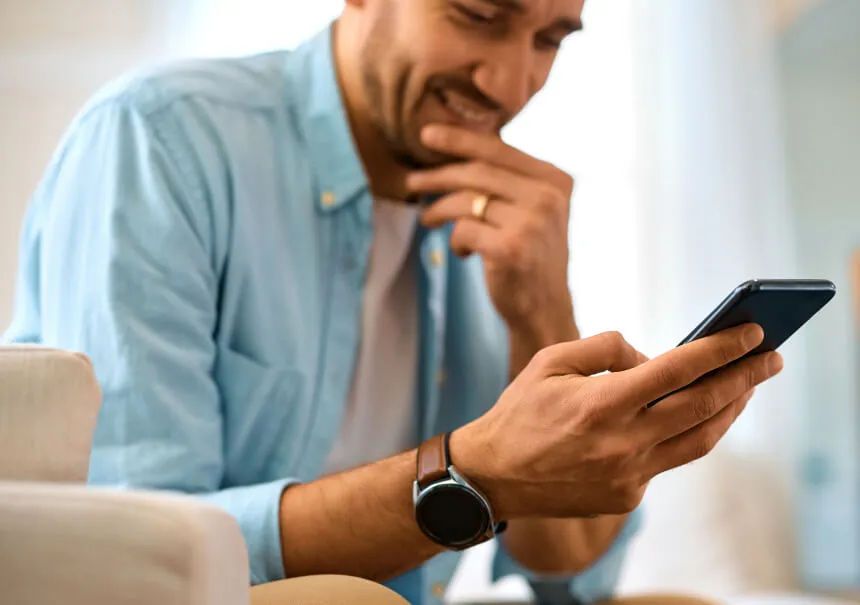 Uśmiechnięty mężczyzna z zaciekawieniem odczytuje spersonalizowane powiadomienie push na ekranie smartfona. Odbiorca w niebieskiej koszuli ze smartwatchem na ręce reaguje na ważny komunikat ze sklepu internetowego.