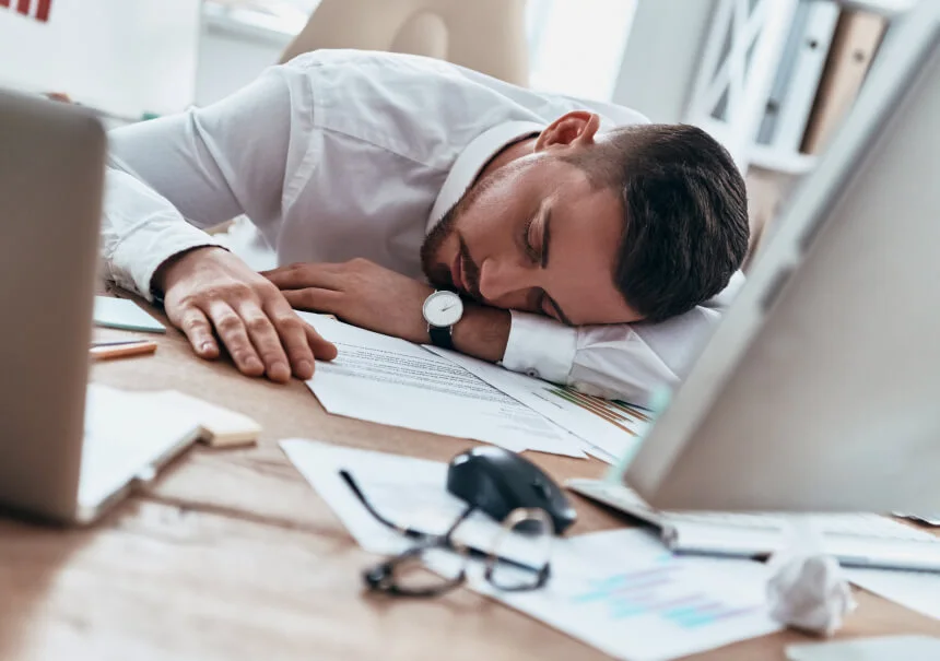 Zmęczony marketer śpi przy biurku pełnym dokumentów. Zamiast ręcznej rutyny, sztuczna inteligencja i automatyzacja marketingu przejmują odtwórcze zadania w firmie.