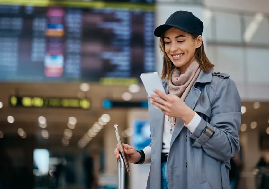 Uśmiechnięta turystka z walizką na lotnisku odczytuje automatyczne powiadomienie WhatsApp przed podróżą z informacjami o rezerwacji.