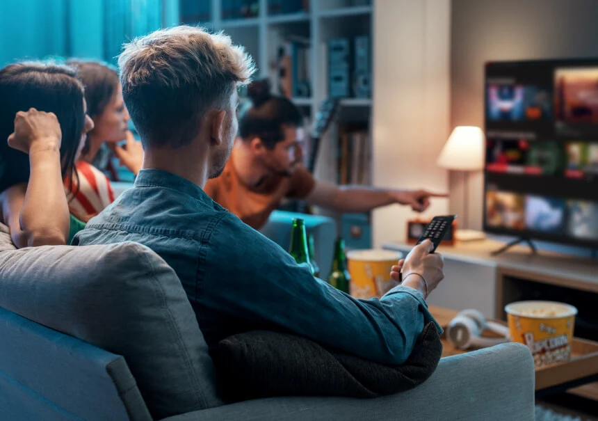 Grupa znajomych ogląda film na platformie VOD, wybierając spersonalizowane rekomendacje treści na ekranie telewizora Smart TV za pomocą pilota.