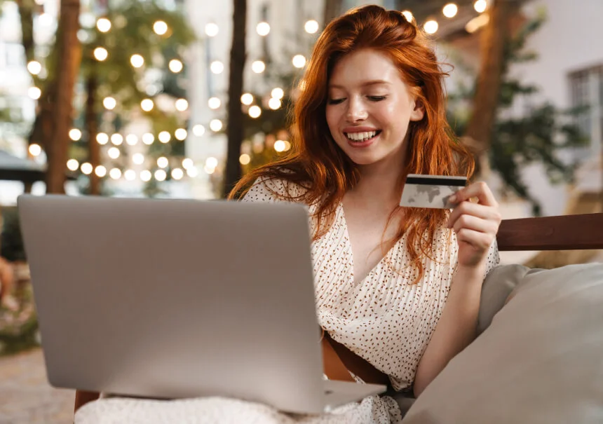 Uśmiechnięta klientka trzyma kartę płatniczą i robi zakupy na laptopie po wykorzystaniu kodu rabatowego z pop-upa w sklepie internetowym.