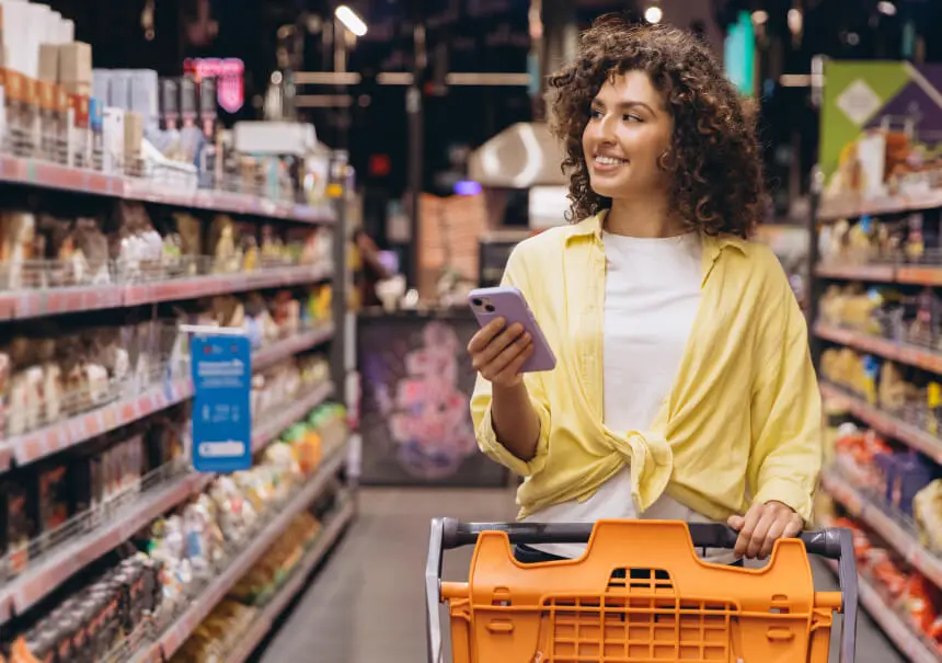 Uśmiechnięta klientka ze smartfonem i wózkiem sklepowym sprawdzająca powiadomienie na WhatsAppie podczas zakupów w markecie FMCG.